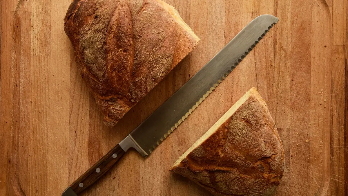 Gekarteld broodmes tussen twee helften van een krokant zuurdesembrood op een houten snijplank