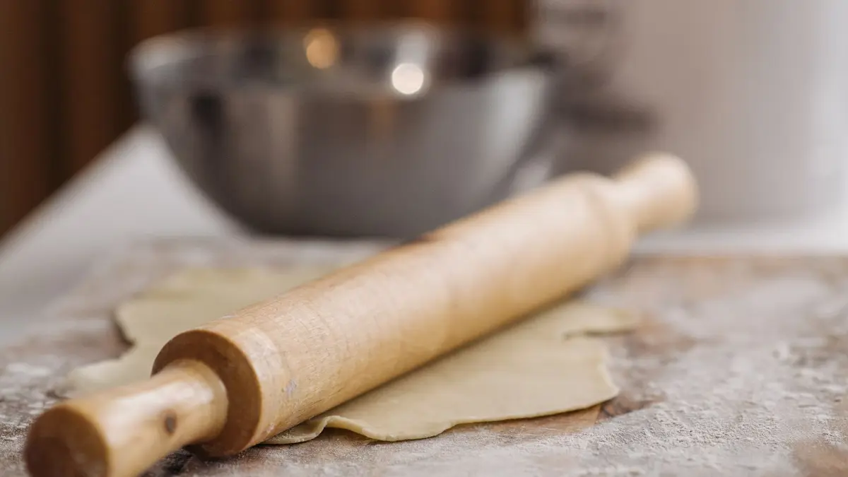 Houten deegroller op uitgerold deeg met een mengkom op de achtergrond