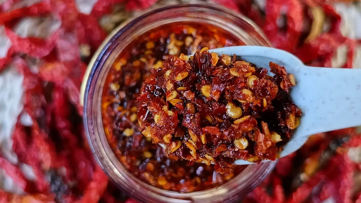 Lepel crispy chili oil boven een pot, omringd door gedroogde chilipepers en knoflook